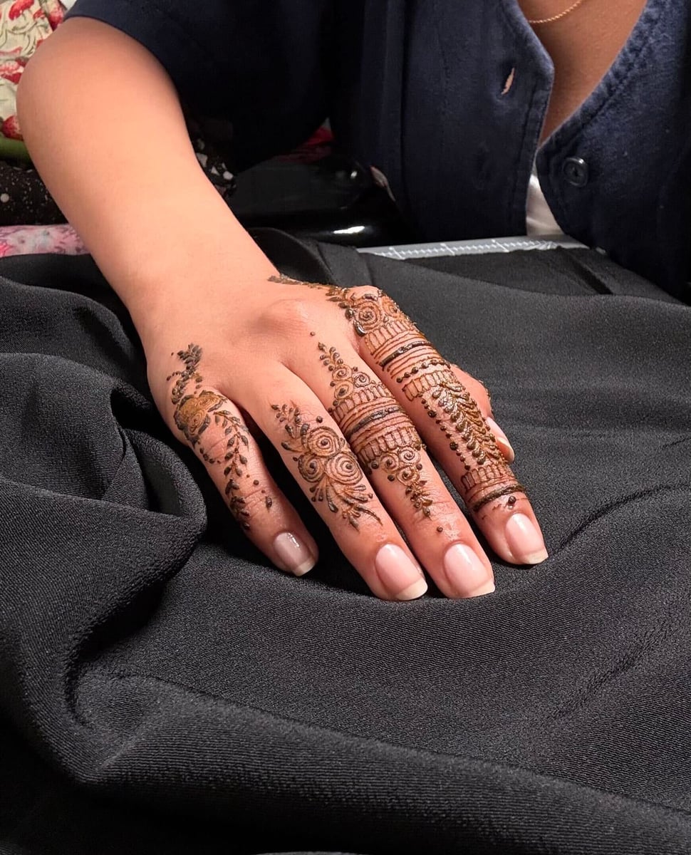 A hand with intricate NooniDesigns henna across the fingers and knuckles, resting on dark fabric.